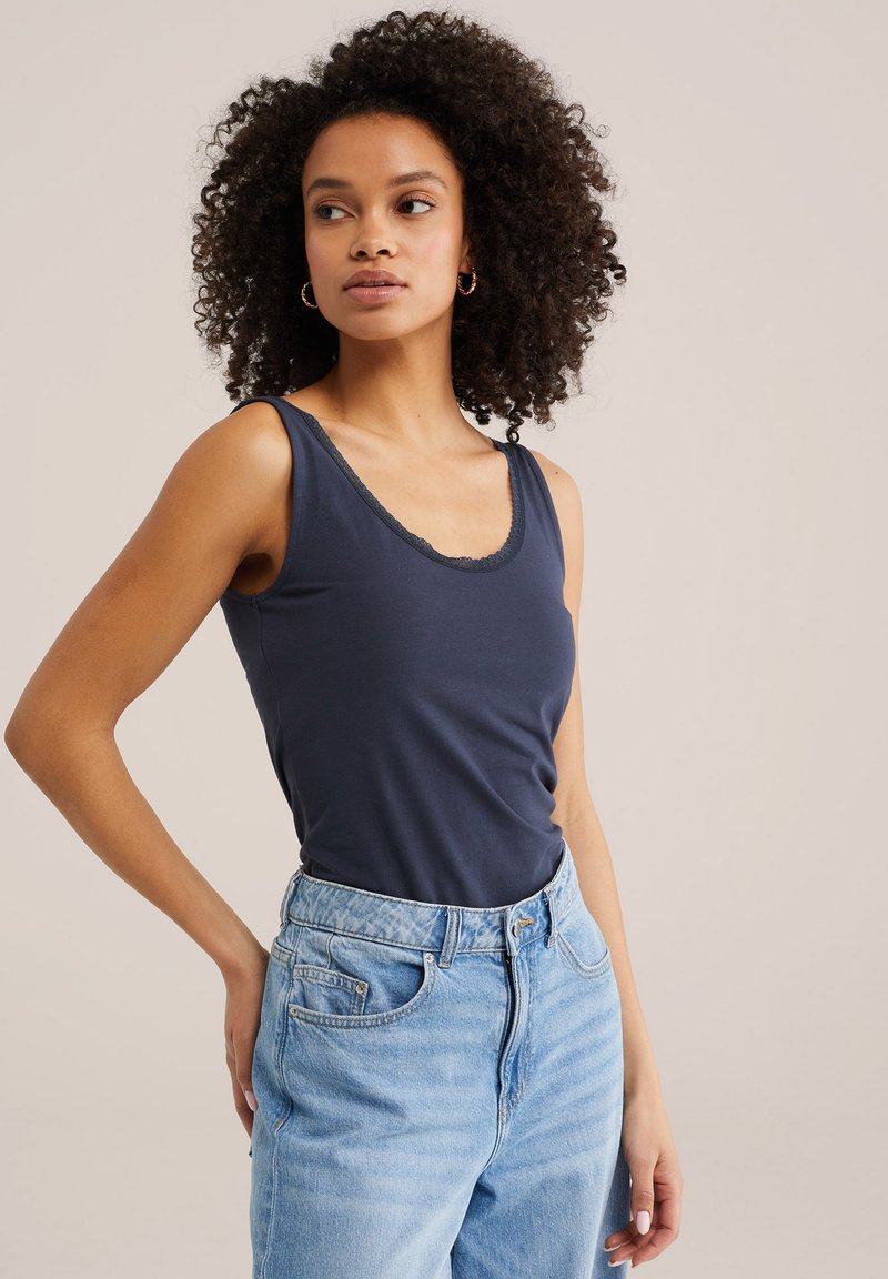 Donna con capelli ricci che indossa orecchini a cerchio dorati, una canotta blu scuro e jeans a vita alta azzurri, posando con una mano sul fianco.