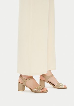 Pieds portant des sandales à talons carrés beiges avec des brides à la cheville, associés à un pantalon long fluide de couleur crème sur un fond clair.