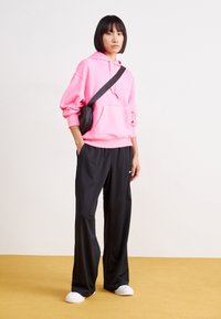 Rosa huvtröja, svarta lösa meshbyxor och vita slip-on skor. Modellen bär en liten svart axelväska. Enkel bakgrund.