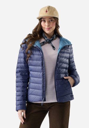 Joven mujer que lleva una chaqueta acolchonada azul, suéter lila claro, pantalones marrones, gorra beige y bufanda negra en el cuello, sonriendo con una mano en el bolsillo.