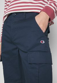 Osoba v námořnických modrých cargo kalhotách s rukou v kapse a červeném a bílém pruhovaném tričku s dlouhým rukávem viditelném od pasu dolů.