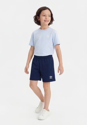 Bambino con capelli corti e scuri che indossa una maglietta Adidas azzurra, pantaloncini Adidas blu navy e sneakers bianche, in piedi e sorridente leggermente di lato.