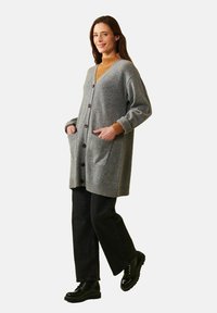 Grå lång kofta med V-ringning, fyra bruna knappar, ribbade muddar och två framfickor, bärs över en orange polotröja och svarta byxor.