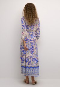 Maxi obleka v modri barvi s vzorcem paisley, cvetličnimi akcenti, dolgimi rokavi in cinčanim pasom, ki se ponaša z večplastnim robom in ravnimi sandalami.