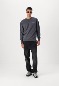 Grå sweatshirt med ribbede mansjetter, som har en liten logo. Brukt sammen med mørke jeans og sølvfargede joggesko. Runde solbriller fullfører stilen.