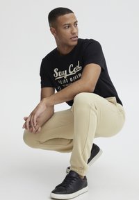 Schwarzes T-Shirt mit dem Aufdruck "Stay Cool", beigefarbene Jogginghose mit elastischen Bündchen und schwarze Sneaker mit weißen Sohlen, in einer Hockposition sitzend.