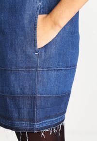 Robe en denim à coupe droite, teinte bleu clair, avec poches latérales et ourlet effiloché. Des coutures visibles créent un motif texturé sur le tissu.