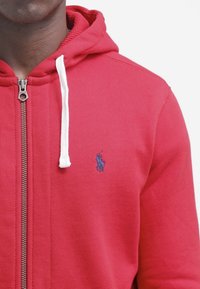 Röd zip-up hoodie i bomullsblandning, med vita dragband, ribbade ärmmuddar och en liten blå hästlogga på vänster bröst.