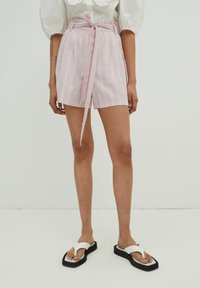 Rosa Leinenbermuda mit weißen vertikalen Streifen, hoher Taille, Bindegürtel und seitlichen Taschen. Kombiniert mit weißen Flip-Flop-Sandalen auf einem schlichten Hintergrund.