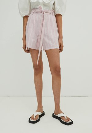 Roze linnen shorts met witte verticale strepen, hoge taille, strikceintuur en zijzakken. Gecombineerd met witte slippers op een effen achtergrond.