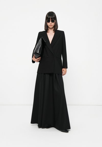 Zwarte op maat gemaakte blazer met een diepe V-hals, gecombineerd met een vloeiende zwarte maxi rok. Het model houdt een zwarte oversized clutch vast en draagt donkere zonnebril.