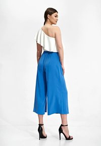 Witte asymmetrische top met ruffles, gecombineerd met blauwe wijde broek. Model draagt zwarte hoge hakken, wat een strakke silhouet benadrukt.