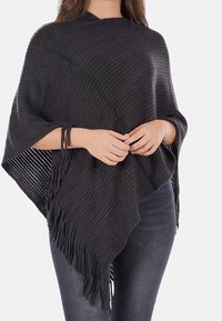 Poncho nero lavorato a maglia con texture a coste, forma triangolare e dettagli frangiati sul fondo, indossato sopra jeans scuri.