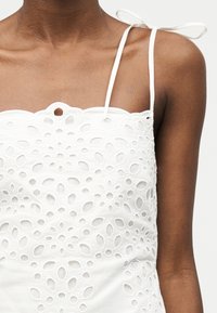 Άτομο με σκούρο δέρμα που φοράει λευκό αμάνικο τοπ με λουλουδάτα σχέδια με μικρές οπές και δεμένες τιράντες στους ώμους.