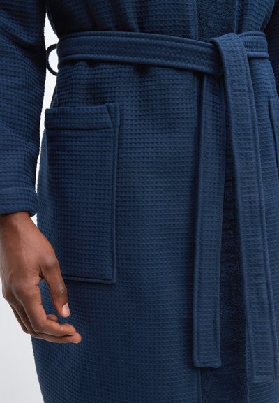 Κοντινό πλάνο ενός ατόμου που φοράει ένα υφασμάτινο ρόμπα σε ναυτικό μπλε, δεμένη στη μέση με ζώνη και με μεγάλο μπροστινό τσέπη.