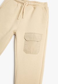 Pantalones de chándal beige con cintura elástica, cordón, bolsillos laterales y un gran bolsillo de parche en el muslo hecho de un material ligeramente diferente.