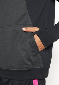 Ręka włożona do kieszeni czarnej i szarej bluzy z kapturem, kciuk przez otwór na kciuk w mankiecie rękawa, nad czarnymi spodniami z różowym paskiem.