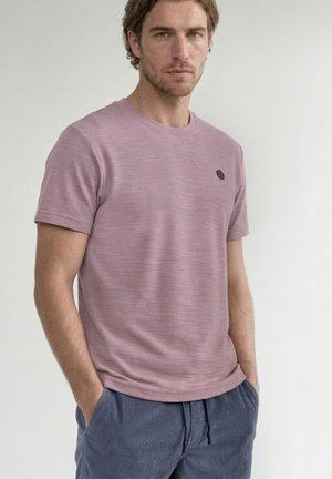 Uomo con capelli castani ondulati, indossa una t-shirt malva a maniche corte e pantaloni blu con coulisse, in piedi con le mani in tasca davanti a uno sfondo neutro.