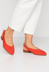 Red suede slingback shoes with a pointed toe and low block heel. Features a small bow detail on the back and smooth texture.