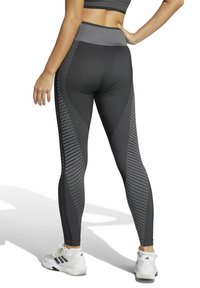 Leggings de patrón negro y gris con una cintura alta, que presentan rayas diagonales y un tejido texturizado, combinadas con zapatillas deportivas blancas.