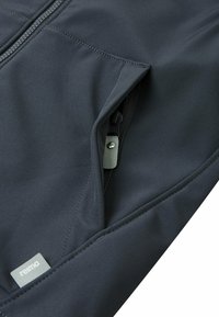 Tissu de veste gris foncé avec une fermeture éclair partiellement ouverte et une petite étiquette affichant la marque "reima". Texture lisse et couture minimale visible.