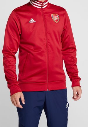Rød lynlåstrøje med hvid Adidas-logo og Arsenal fodboldklub-logo, båret med marineblå Adidas træningsbukser.