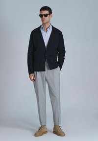 Blazer en tricot marine avec deux poches avant, chemise bleu clair en dessous, pantalon ajusté gris clair et chaussures en suède beige avec lacets.