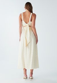 Žena nosi bezrukavnu krem midi haljinu s crnim točkicama, otvorenim leđima koja je vezana mašnom, i bijelim sandalama s petom.