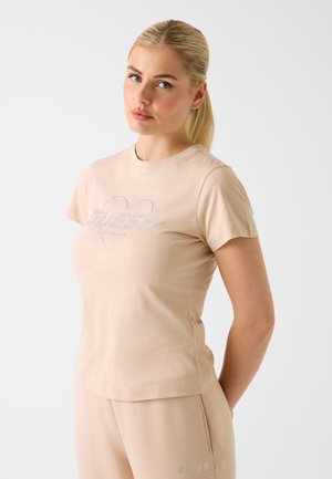 Blonde woman wearing matching beige Guess t-shirt and pants, standing with hands behind her back against plain white background.