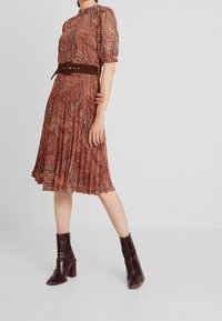 Rode en oranje gestreepte jurk met korte pofmouwen, een hoge halslijn en een bruine riem; gecombineerd met donkerbruine enkellaarzen.