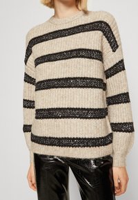 Beige tröja med svarta texturerade ränder, rund halsringning och långa ärmar; bärs med glänsande svarta byxor. Mjuk stickad material.