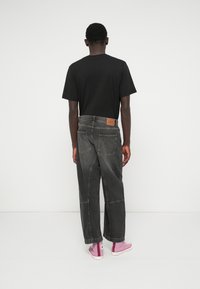 Άνδρας στέκεται με την πλάτη γυρισμένη, φορώντας μαύρο μπλουζάκι, χαλαρούς σκούρους γκρι τζιν κι ροζ αθλητικά παπούτσια ψηλής �φτέρνας, απέναντι από ένα απλό ανοιχτό φόντο.