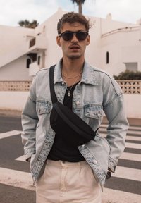 Džínová bunda ve světle modré, černá kabelka přes rameno, vrstvené stříbrné náhrdelníky, černé tričko, světle krémové kalhoty, modelováno v městském prostředí.