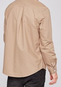 Beige overhemd met lange mouwen, een afgeronde zoom, een plooi aan de achterkant en manchetten met knopen. Soepele katoenen stof, zonder patronen of afbeeldingen.