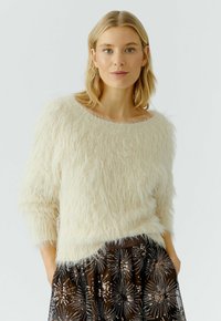 Blonde Frau, die einen cremefarbenen, flauschigen Pullover und einen dunklen Rock mit silbernen und bronzenen Pailletten-Sternmuster trägt, mit den Händen in den Taschen.