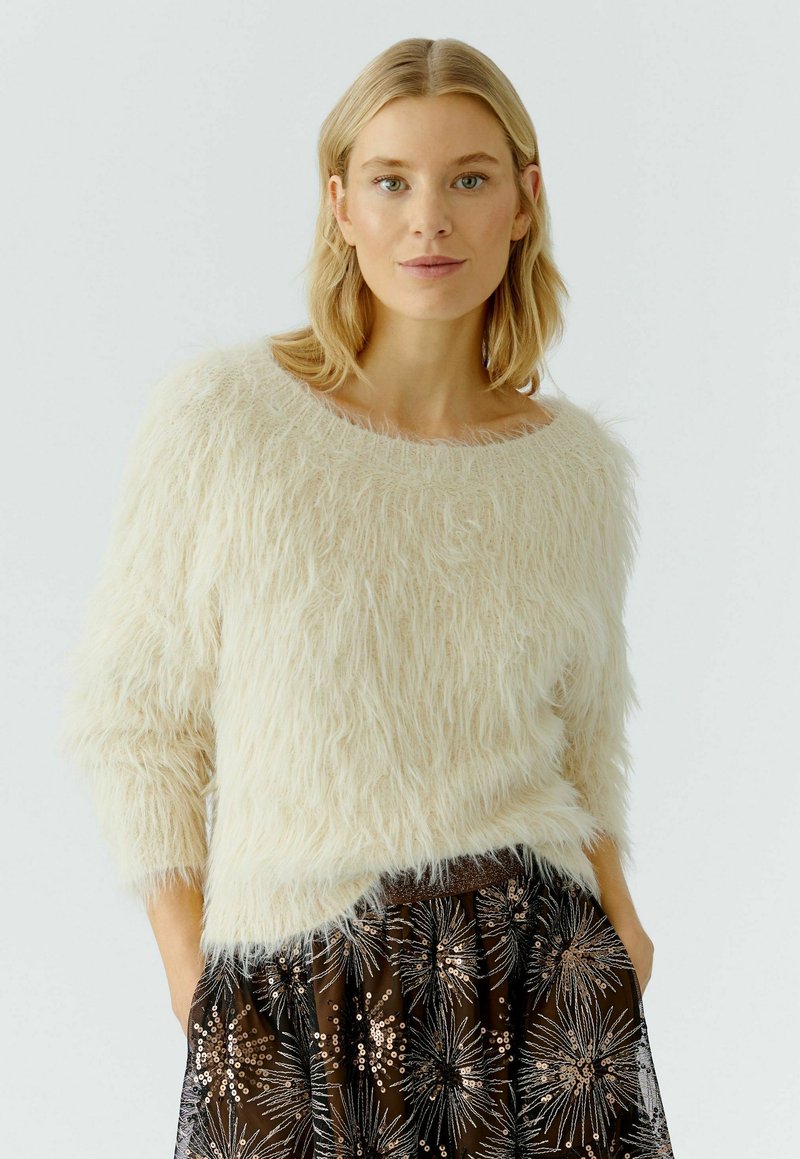 Blonde Frau, die einen cremefarbenen, flauschigen Pullover und einen dunklen Rock mit silbernen und bronzenen Pailletten-Sternmuster trägt, mit den Händen in den Taschen.