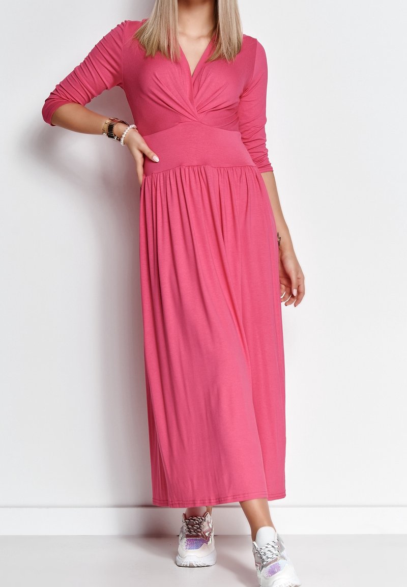 Femme portant une longue robe rose à manches longues et des baskets blanches, debout avec une main sur la hanche, sur fond blanc.