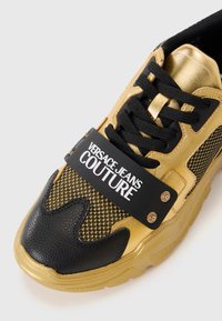 Guld- og sort sneakers med mesh- og læderteksturer, prydet med en mærkesnor og kontrastfarvede detaljer. Kraftig sål og snørebåndslukning.