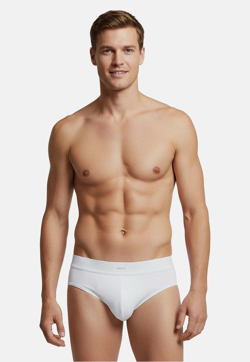 Homme en forme debout, portant un slip blanc, souriant légèrement, sur un fond gris clair uni.