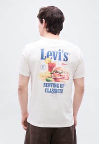 Άνδρας που φοράει λευκό t-shirt Levi's με σχέδιο μπέργκερ, πατάτες τηγανητές, ποτό και το κείμενο «Serving Up Classics! Since 1873» στην πλάτη.