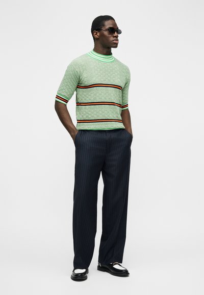 Muž nosiaci čierne slnečné okuliare, svetlozelený krátky sveter s textúrou a oranžovými pruhmi, námornícke nohavice s tenkými prúžkami a čierno-biele topánky.