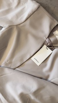 Blusa de tela suave y gris claro, con cuello redondo, costuras en los hombros y etiquetas de cuidado adjuntas. Presenta un diseño sencillo y sin patrones visibles.