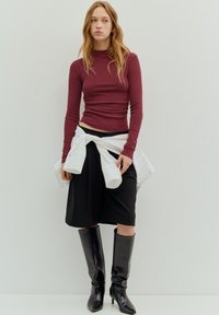 Bordeauxrotes Langarmoberteil, schwarzer Rock und kniehohe schwarze Stiefel mit Absatz. Weißes Hemd um die Taille gebunden. Schlichtes, figurbetontes Design.