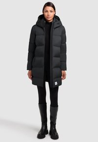 Black puffer coat with a hood, featuring a zip front, large quilted sections, and a small logo patch at the hem. Worn over black leggings and boots.