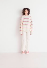 Haut à manches longues de couleur crème avec des rayures roses et vertes, associé à un pantalon blanc évasé et des baskets roses. Cheveux bouclés encadrant le visage.