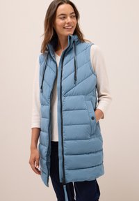 Femme souriante portant une veste sans manches en quilt bleu clair sur un haut à manches longues blanc, avec une main dans la poche de la veste, fond beige.