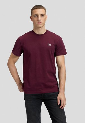 Hombre de pie de frente, lleva una camiseta Lee burdeos y vaqueros oscuros sobre un fondo liso gris claro.