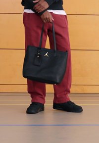 Svart lærveskepose med doble håndtak, gull logo detalj og inkludert merkelapp. Brukt med røde bukser og svarte sko. Overflaten er glatt.