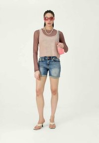 Top cropped rosa claro sobre uma camisa de manga longa castanha, combinado com calções de ganga e sandálias brancas. Os acessórios incluem óculos de sol e uma bolsa rosa.