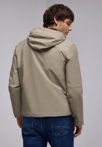 Veste beige résistante à l'eau avec capuche, présentant une texture lisse et un design ajusté, associée à un jean en denim bleu foncé.